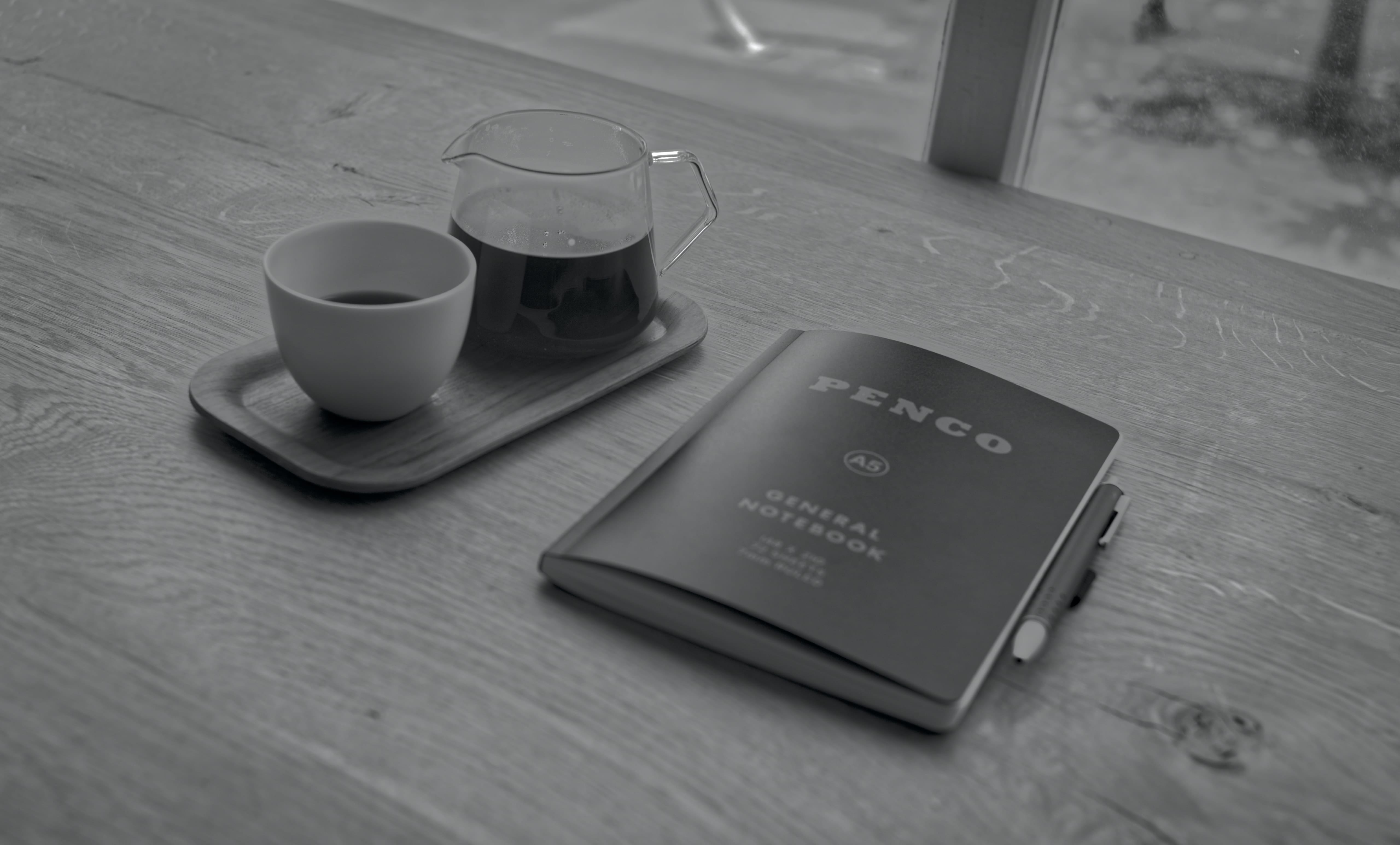 Black-and-white photograph of a coffee carafe and cup beside a Penco notebook