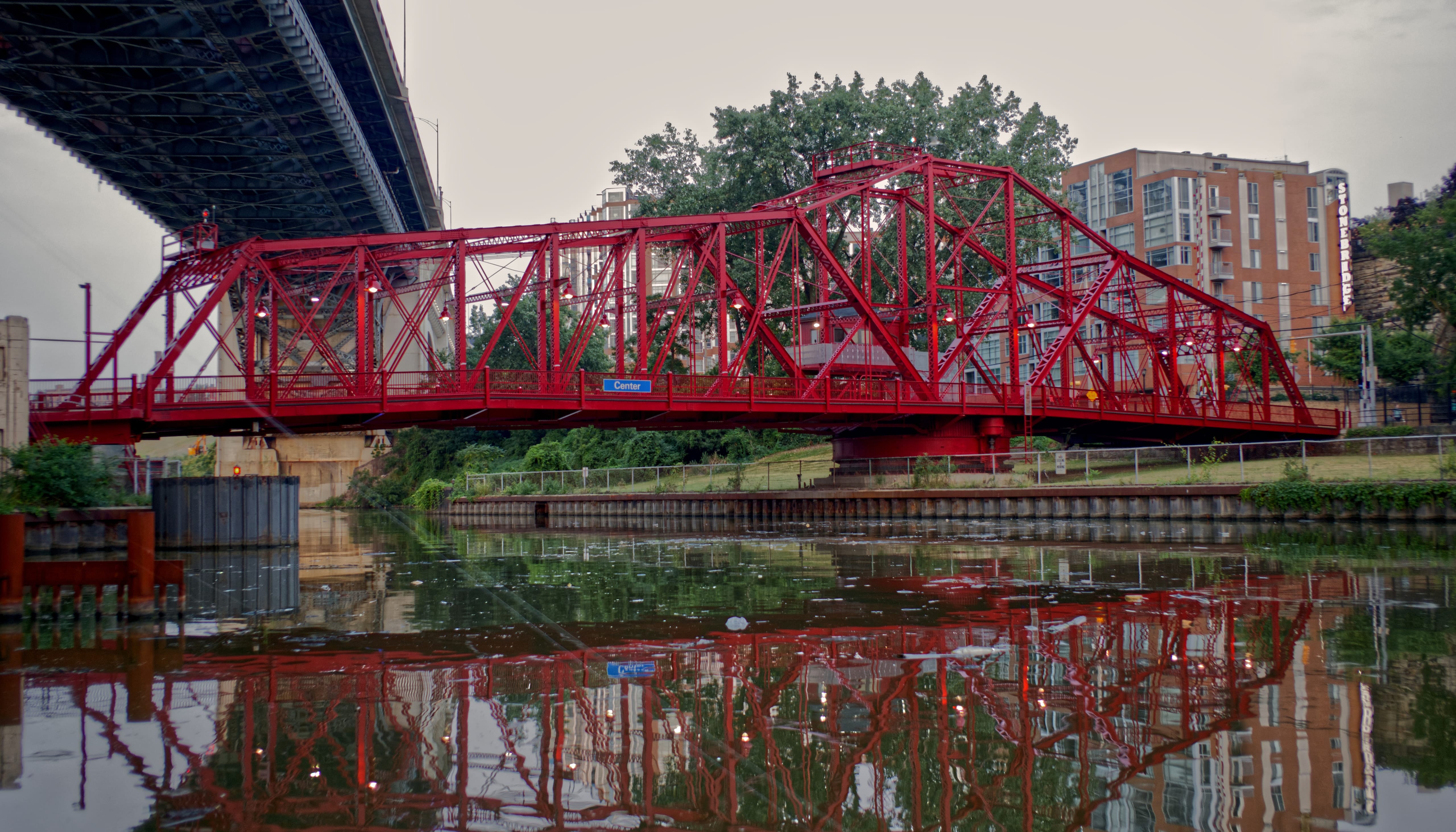 A red steel swing bridge crossing the Cuyahoga River, reflected in the water beneath a larger bridge