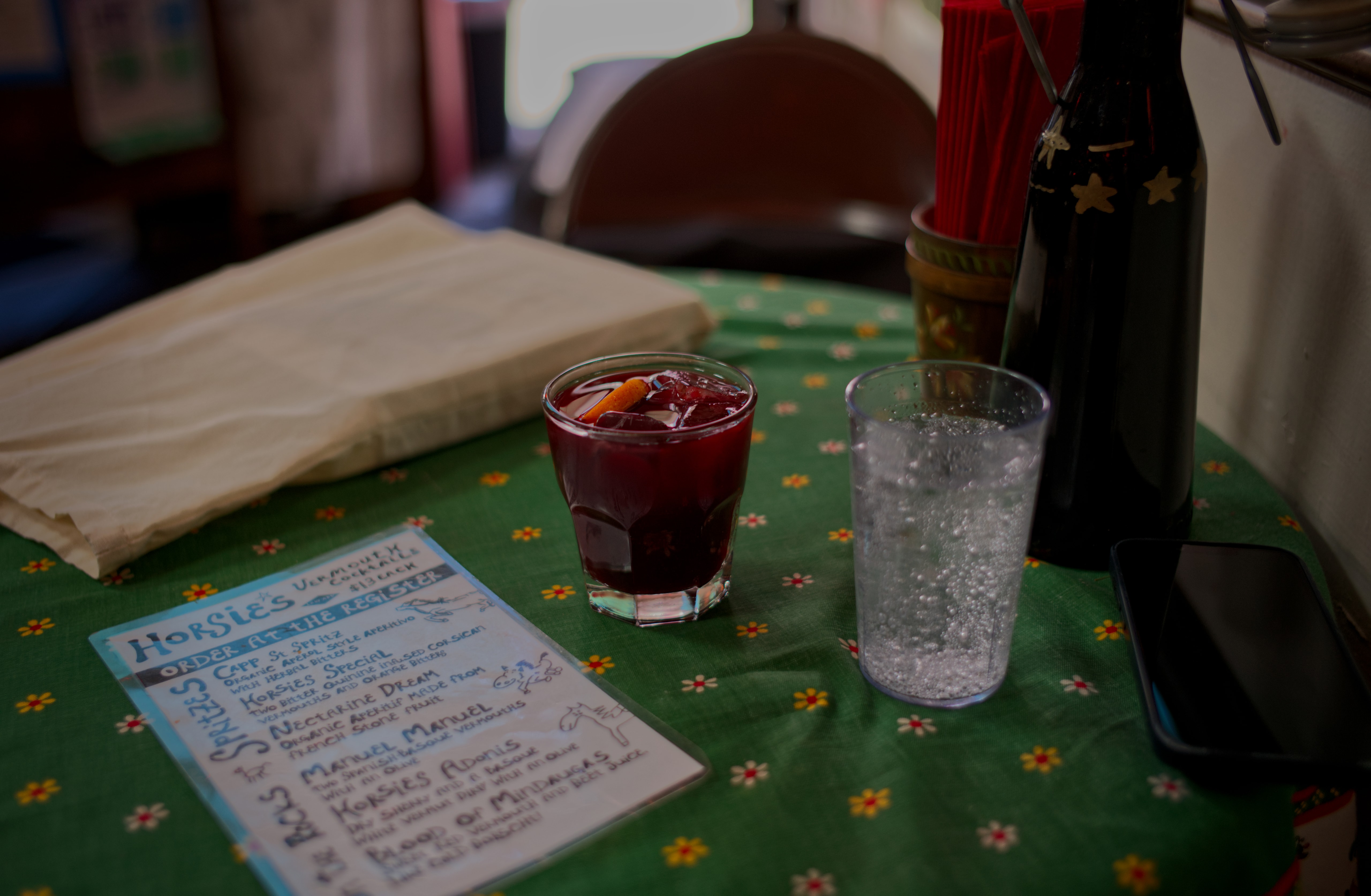 A red Borscht cocktail with an orange peel on a floral tablecloth beside a glass of water and a drink menu