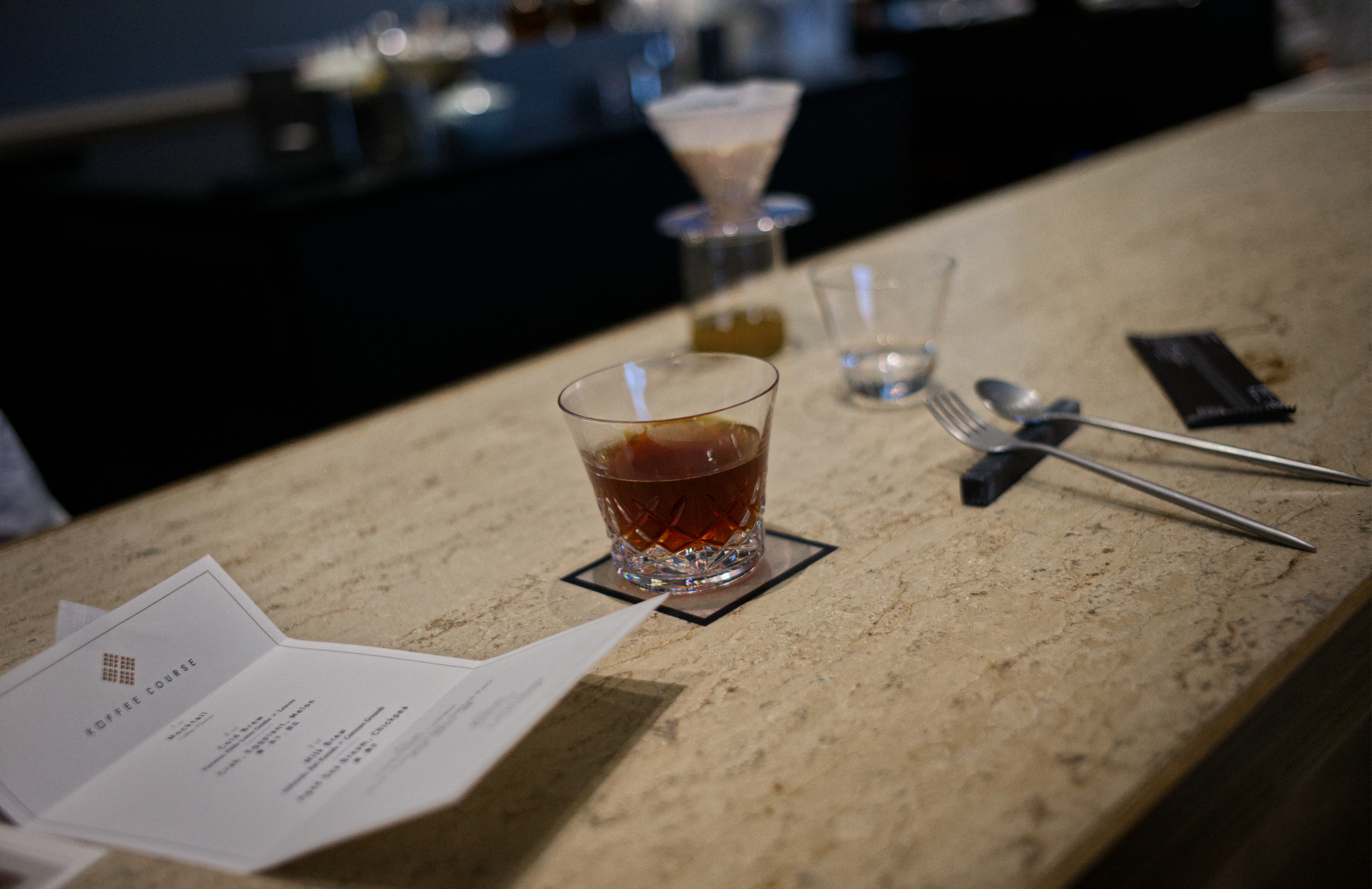 A dark coffee drink in a low glass beside a folded Koffee Course menu at Koffee Mameya Kakeru